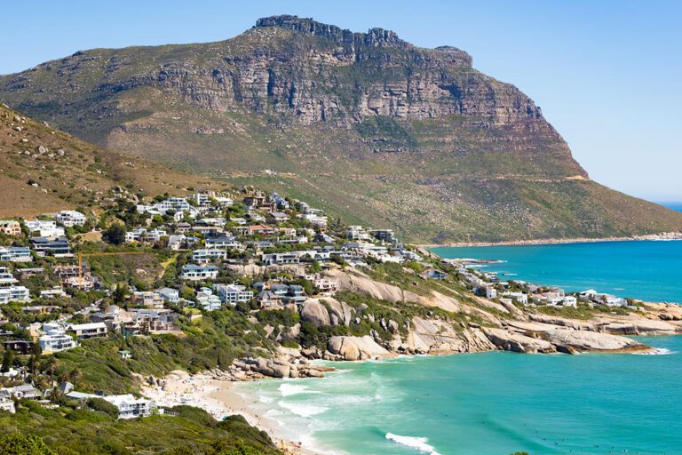 Beautiful coastal landscape of Cape Town, featuring a built-up residential area on a steep slope overlooking a pristine beach with turquoise waters and a towering mountain backdrop.