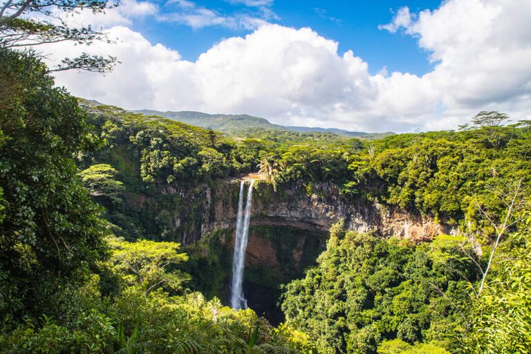 A beautiful natural scenery shot of Chamarel Falls, highlighting the tall, slender waterfall amidst vibrant green vegetation and rolling hills, with dramatic clouds overhead.