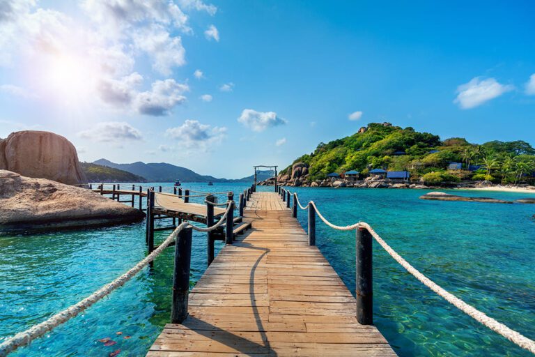 The iconic wooden bridge connecting parts of Koh Nangyuan Island in Thailand, showing clear calm waters, distant hills, and a sunny tropical atmosphere.