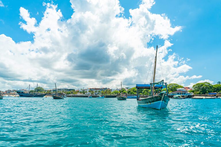 Traditional Dhow boats anchored in the turquoise waters off the coast of Stone Town, Zanzibar, with the historic city skyline visible under a bright blue sky with large white clouds.